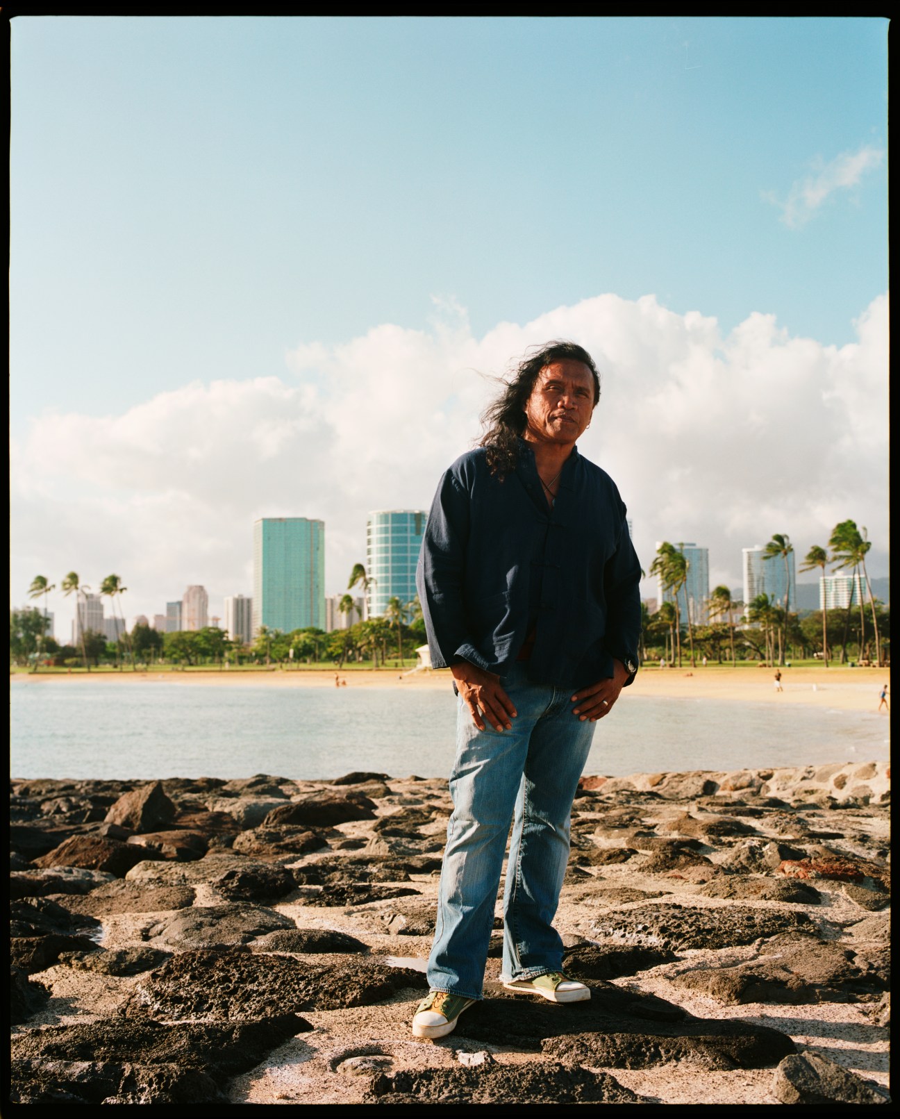Henry Kapono, musician and activist, stands on a rocky beach with Waikiki skyline and palm trees behind him.