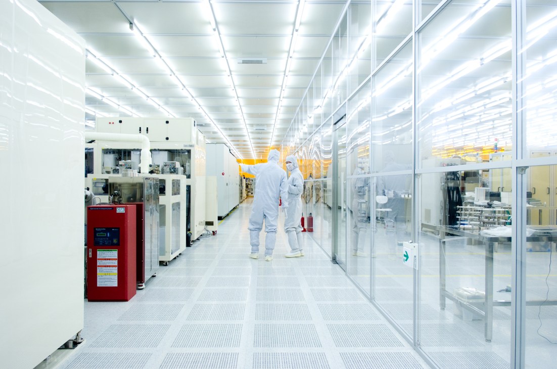 Two technicians in protective white suits work in a pristine clean room facility with advanced semiconductor manufacturing equipment lining both sides.