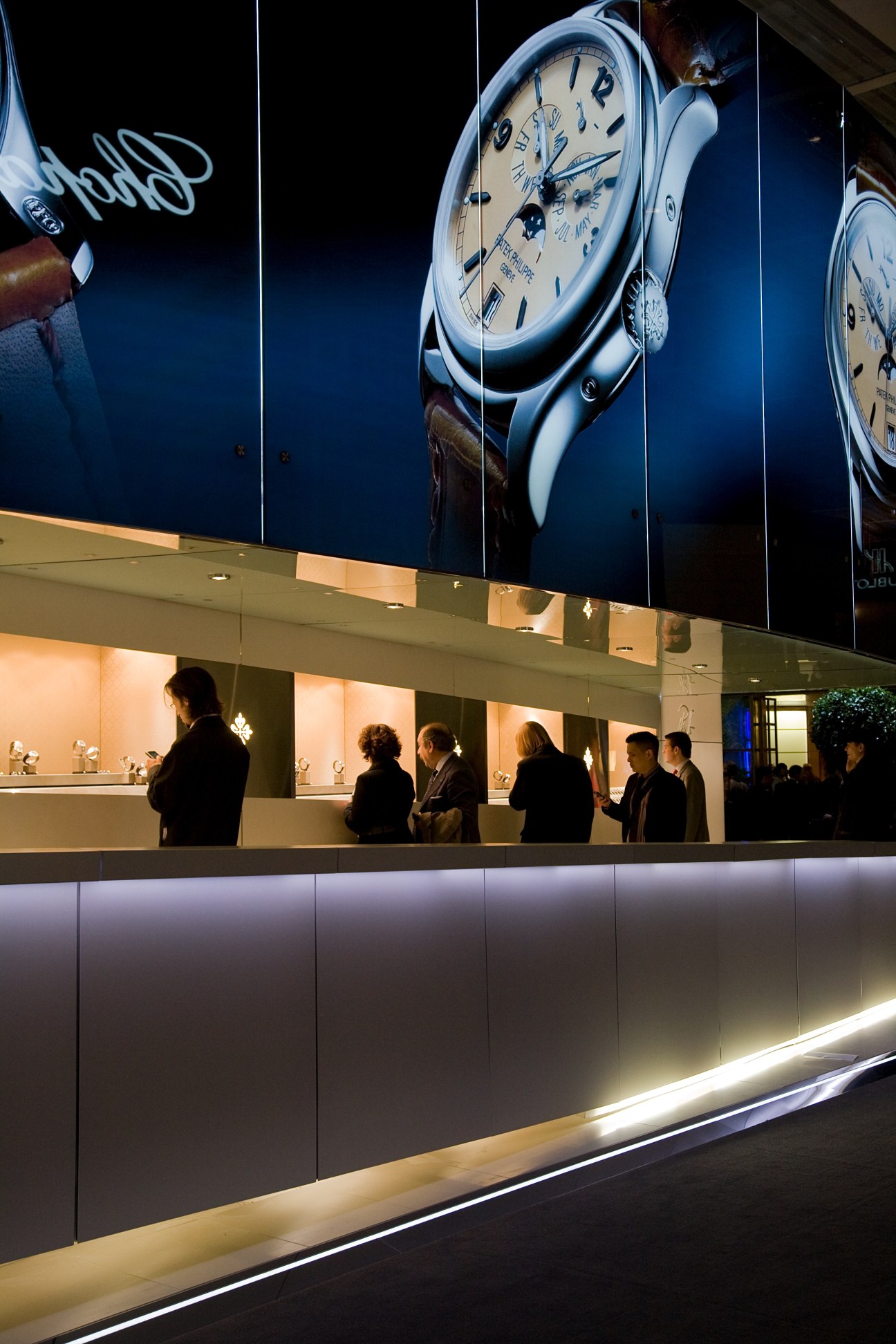 Visitors browse watches at a Patek Philippe exhibition stand with large illuminated watch displays above.