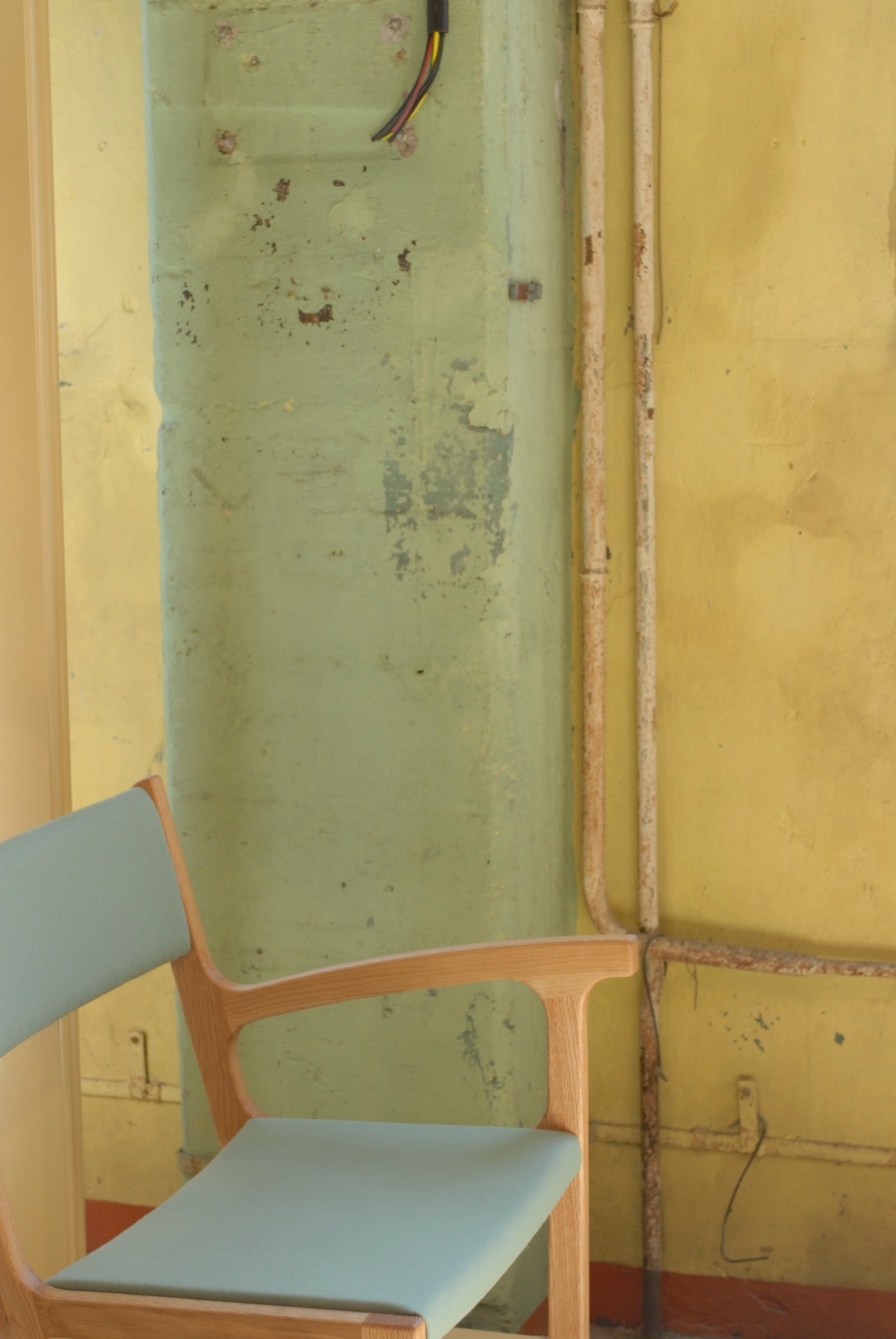 One-armed chair by Autoban for De La Espada with wooden frame and light blue seat against weathered green and yellow wall.