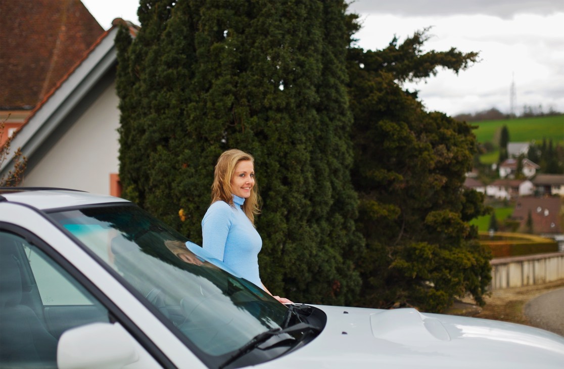 Sandra Huber stands beside a white Subaru Forester turbo in a rural Swiss setting with hedges and farmland behind her.