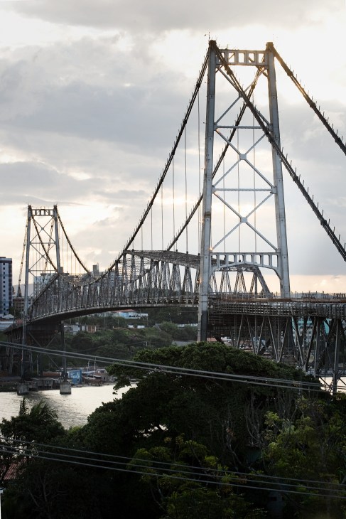 The Hercílio Luz suspension bridge spans across water in Florianópolis, Brazil, with steel cables and structural supports visible against a cloudy sky.