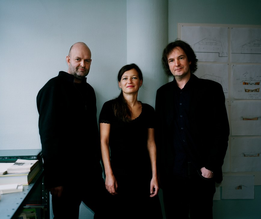 Three architects in black clothing standing in an office with architectural drawings on the wall behind them.