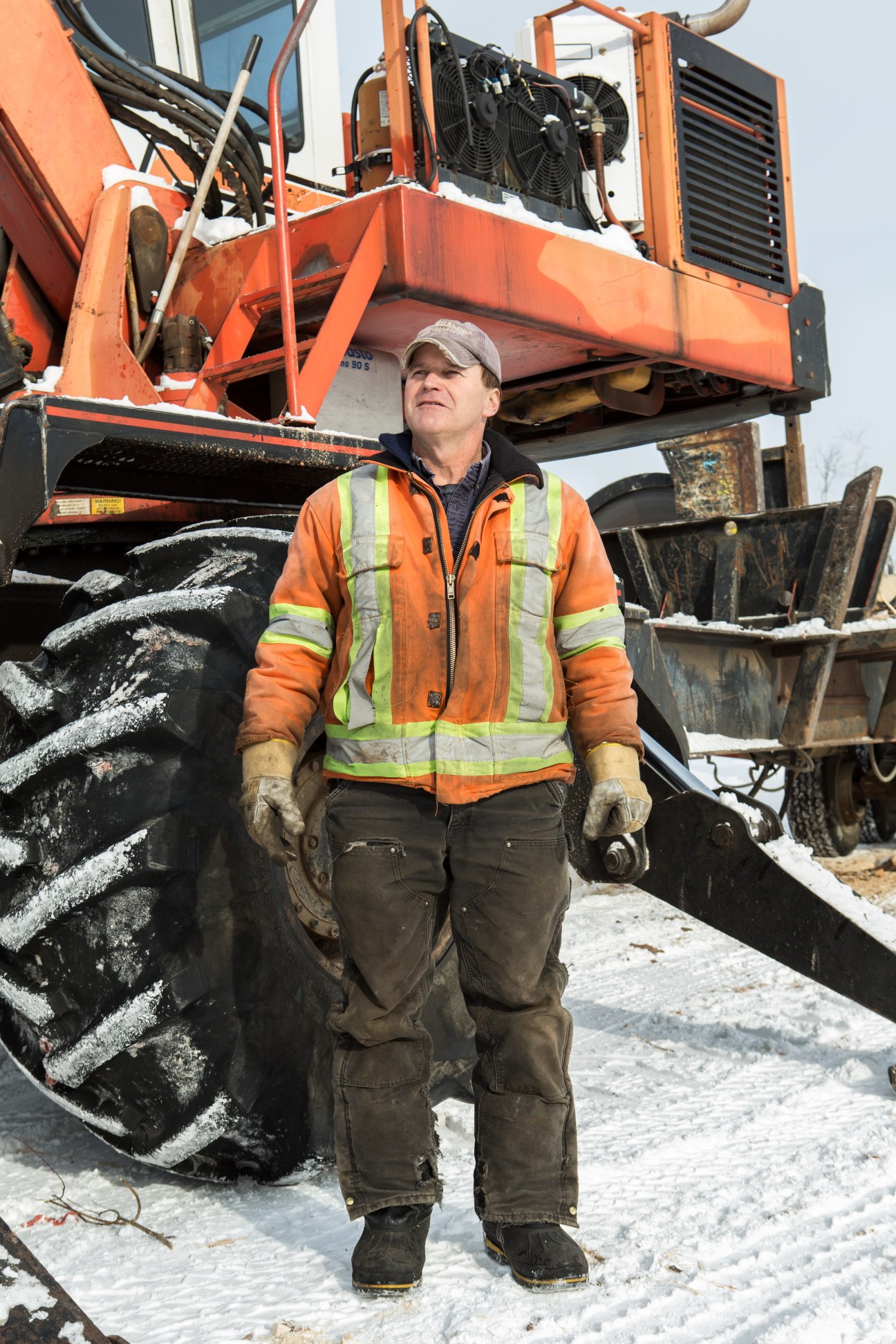 Man in orange safety vest and cap stands in front of large red forestry equipment in snowy conditions.