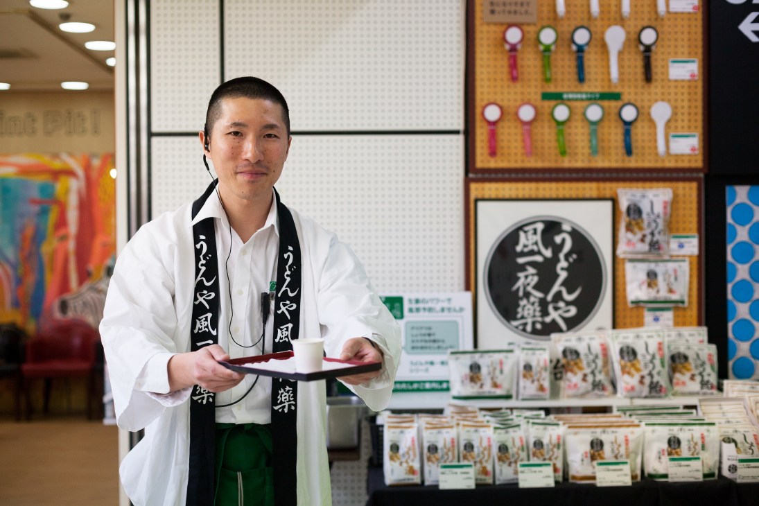 Shintaro Watanabe, a Tokyu Hands staff member wearing a white shirt and black scarf, stands in front of a display of products and pegboard shelving.