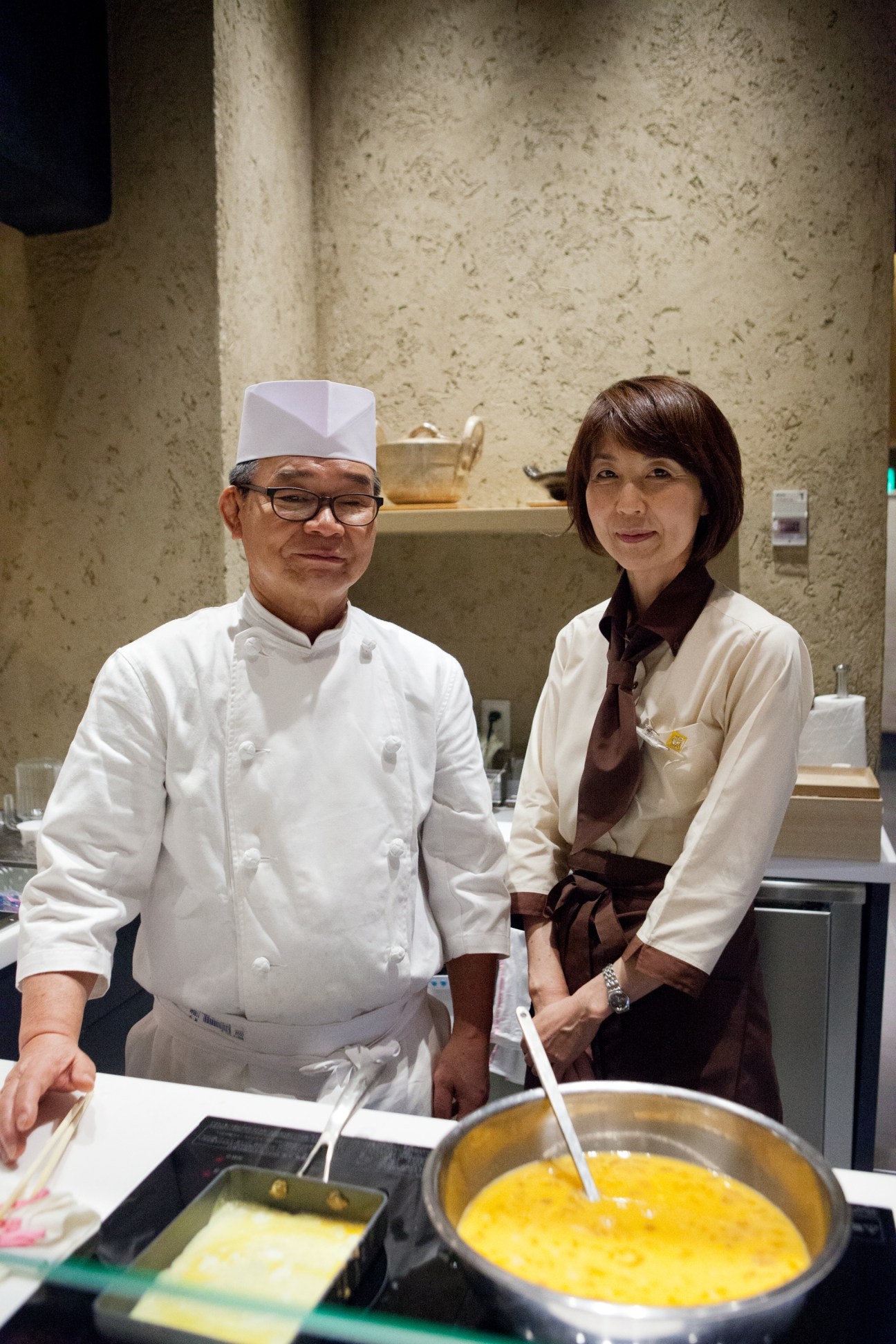 A chef in white uniform and hat stands with a woman in cream and brown clothing at Kayanoya restaurant, with a pot of soup in the foreground.