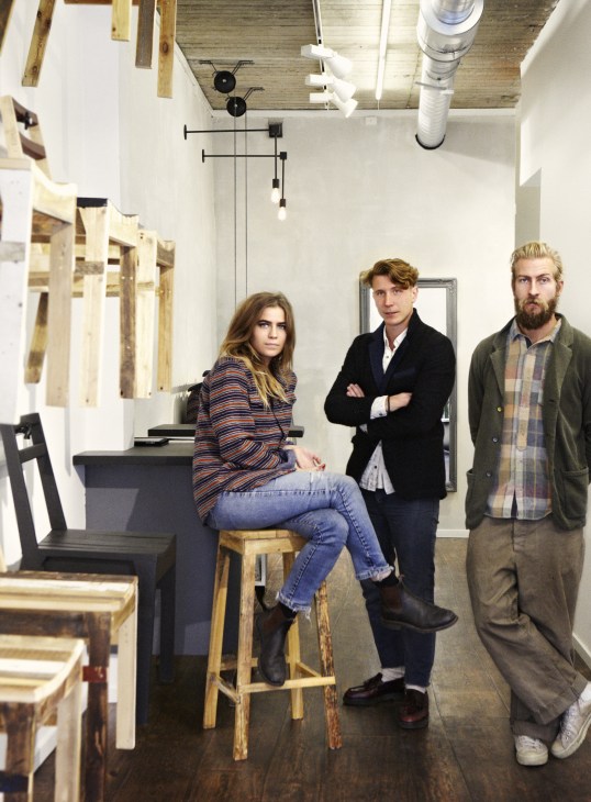 Three people pose in a minimalist retail space with wooden elements and pendant lighting.