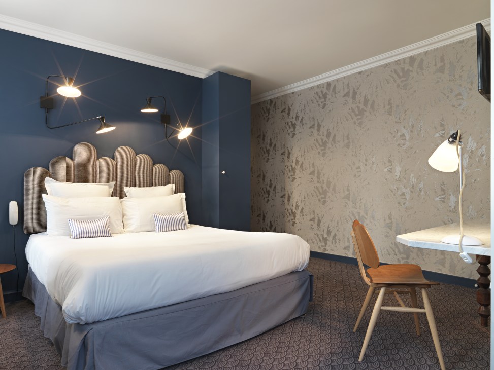 Hotel Paradis bedroom with blue accent wall, ribbed headboard, white bedding, and mid-century modern furnishings.