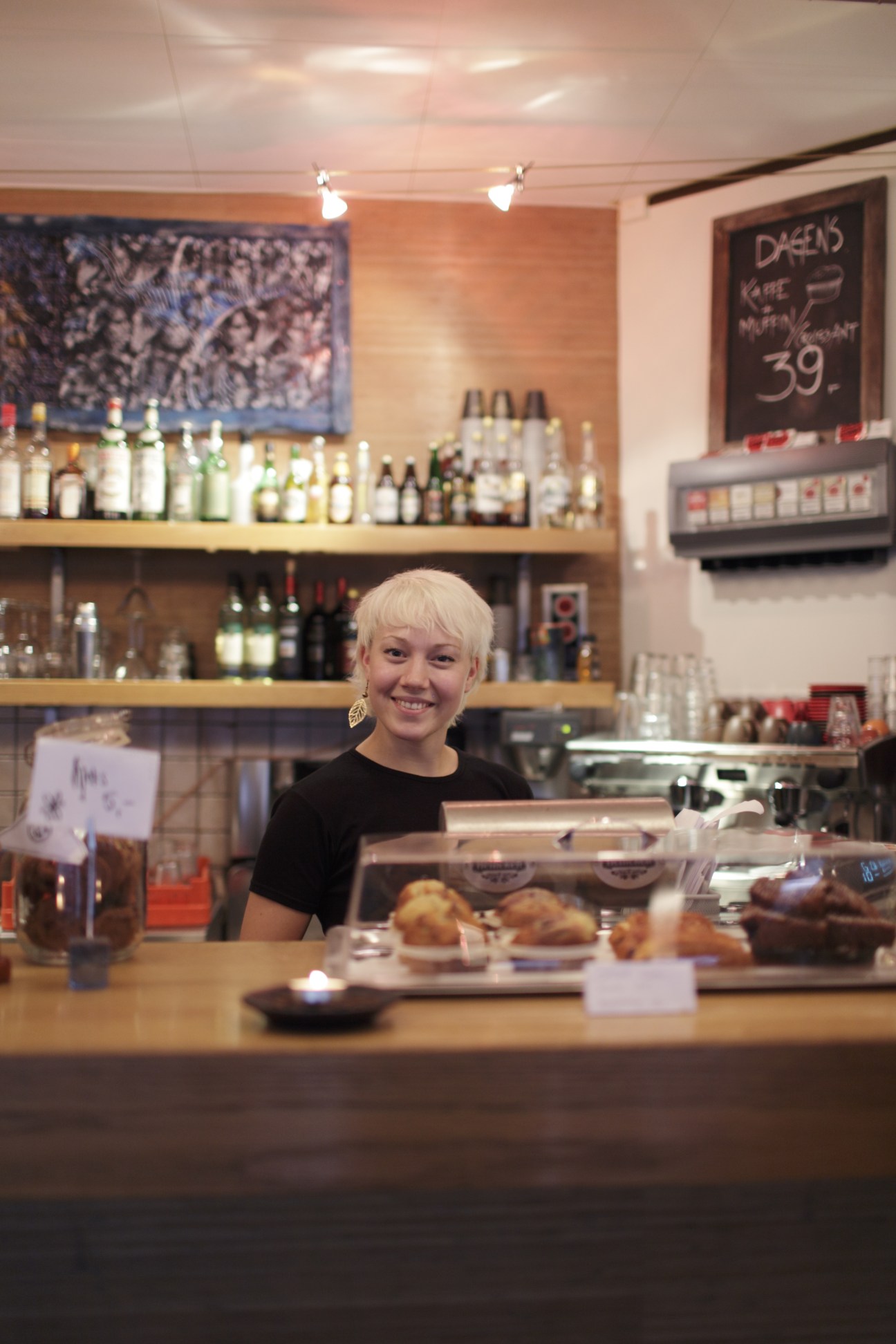 Åse Fredheim, breakfast waiter at Nero café, smiles behind the counter with shelves of bottles and a menu board behind her.