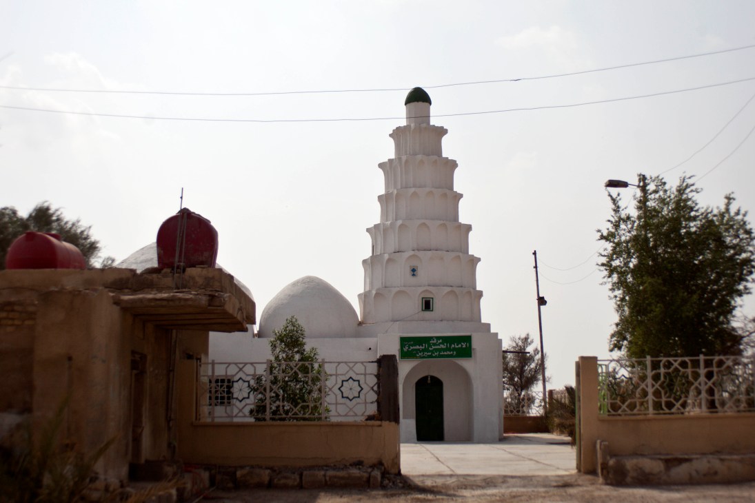White mosque with minaret tower and red dome, ornate geometric gate, palm trees visible in Basra.