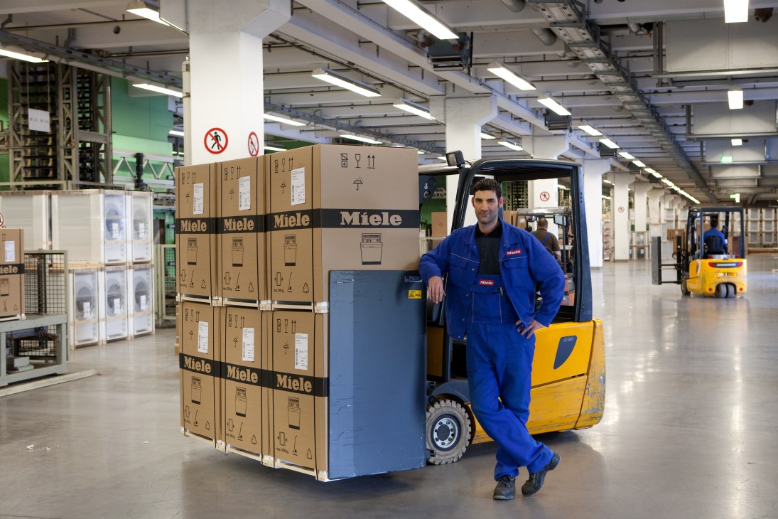 A Miele factory worker in blue uniform poses with a forklift loaded with cardboard boxes of dishwashers in the Gütersloh manufacturing plant.