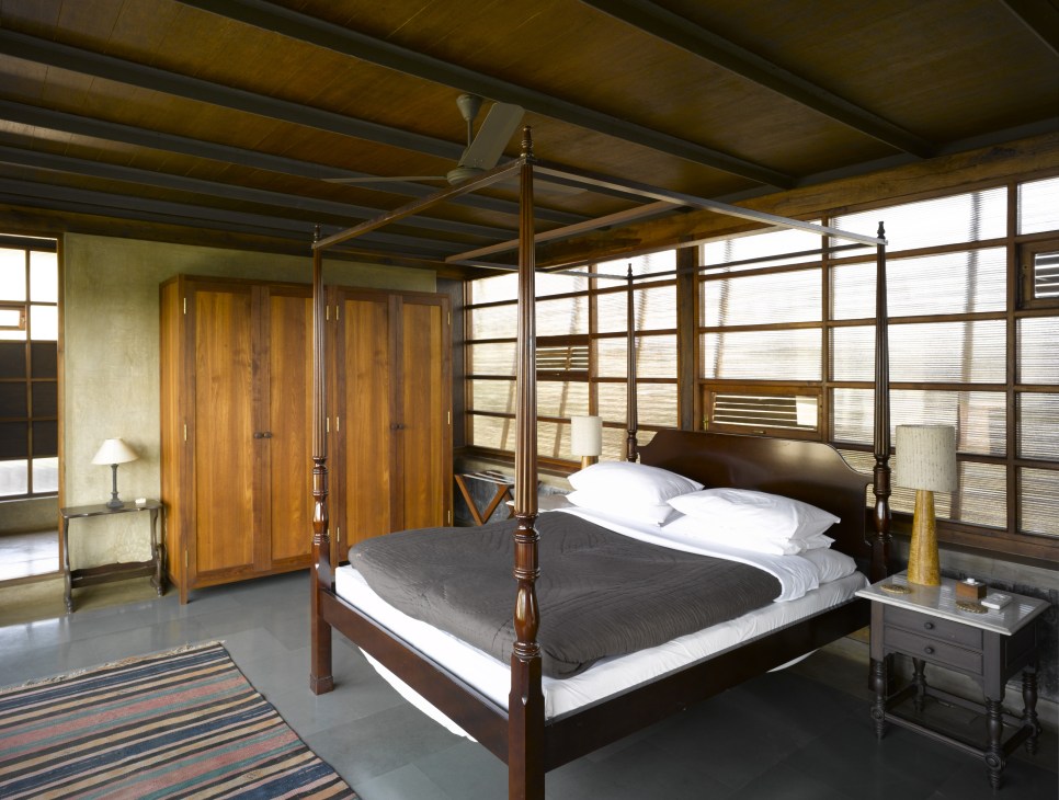 Spacious bedroom with four-poster bed, wooden wardrobe, slatted screens filtering light, and ochre-toned walls.