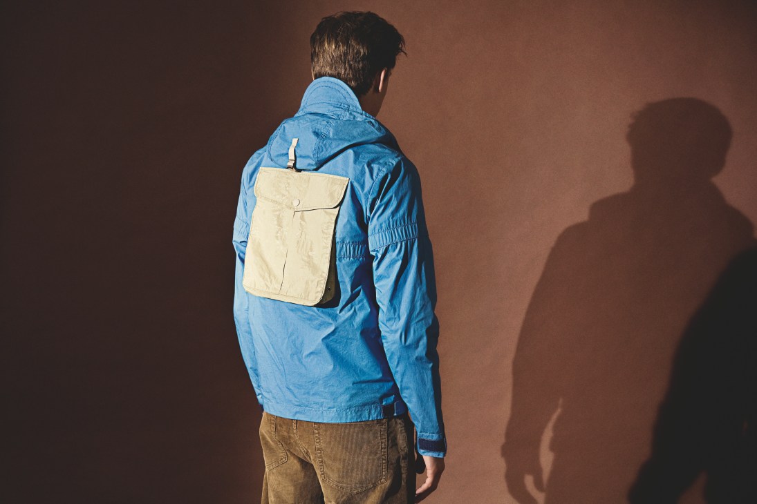 Person wearing a blue denim jacket with a cream canvas backpack against a brown wall.