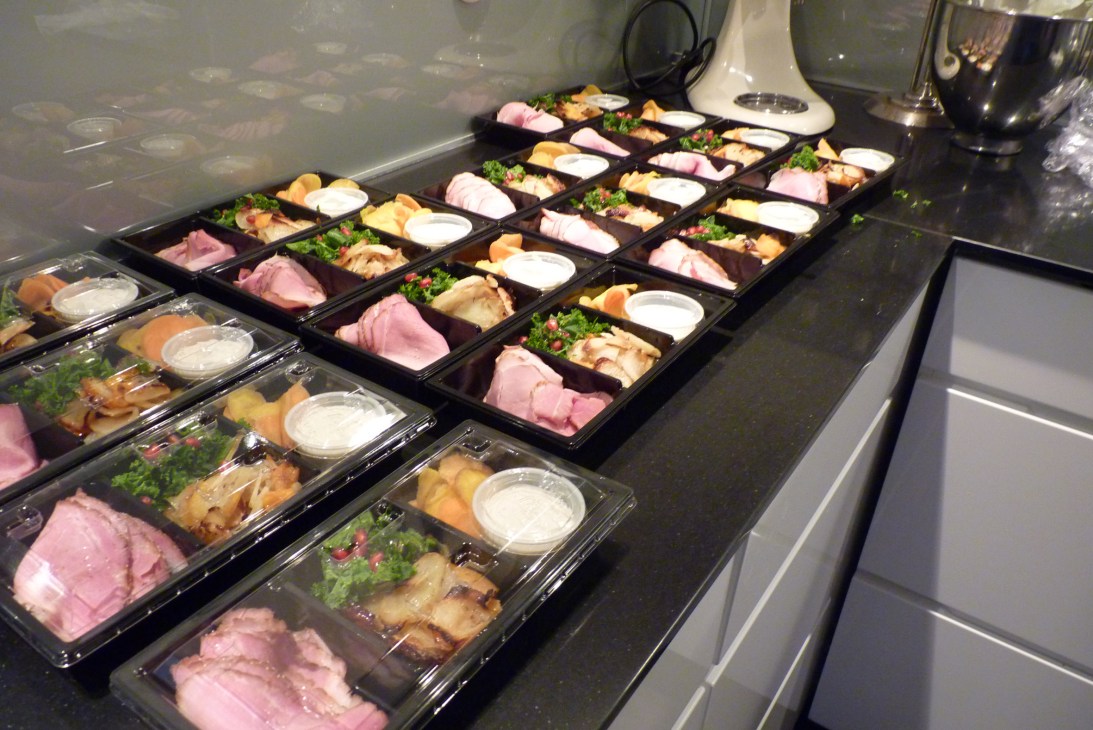 Multiple black trays of prepared airline meals with ham, chicken, pastries, greens and sour cream portions.
