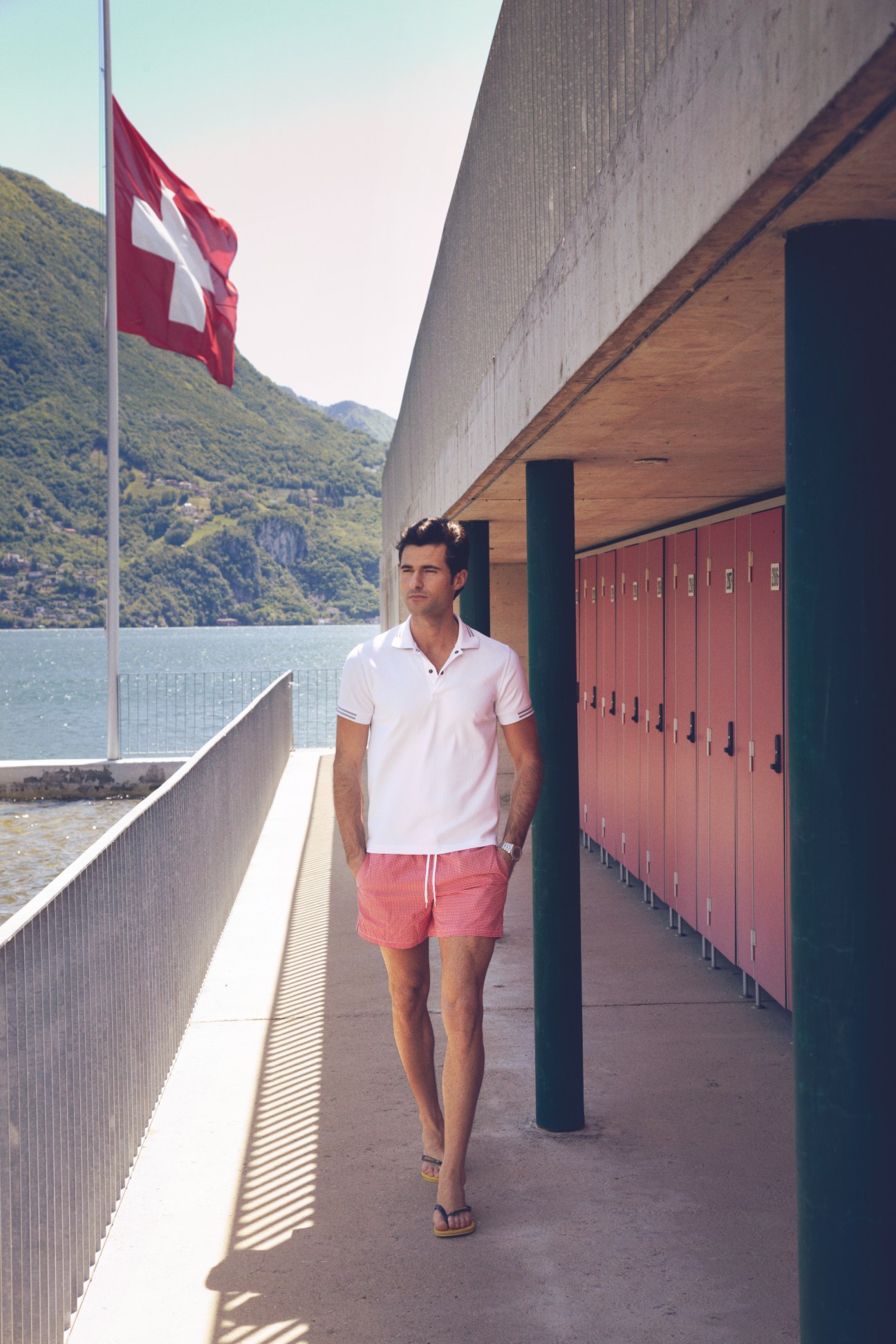 Man in white polo shirt and coral swim trunks walks along a lakeside promenade with Swiss flag and mountains in background.