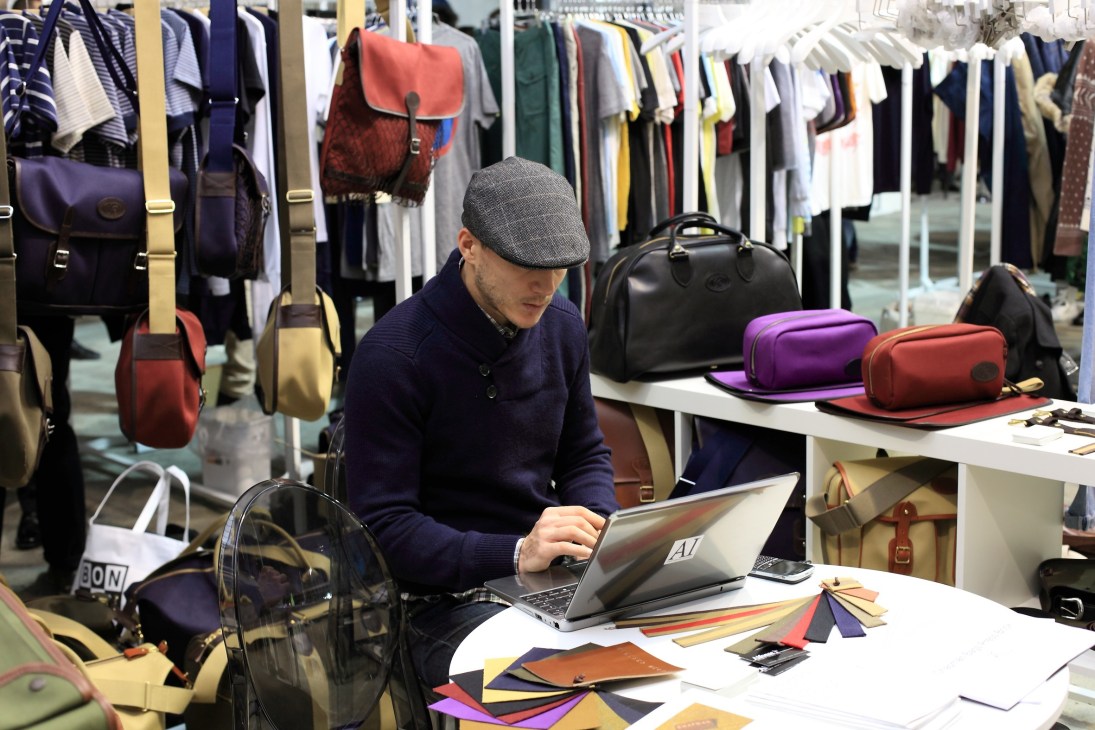 A man in a navy sweater and flat cap sits at a trade fair booth working on a laptop, surrounded by leather goods and clothing displayed on racks.