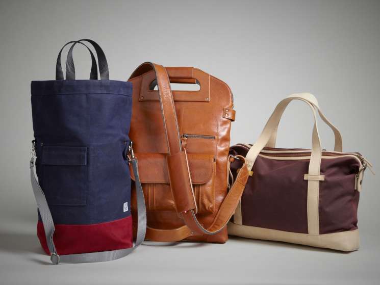 Three bags displayed: navy and red tote, cognac leather backpack, and burgundy with cream handles shoulder bag.