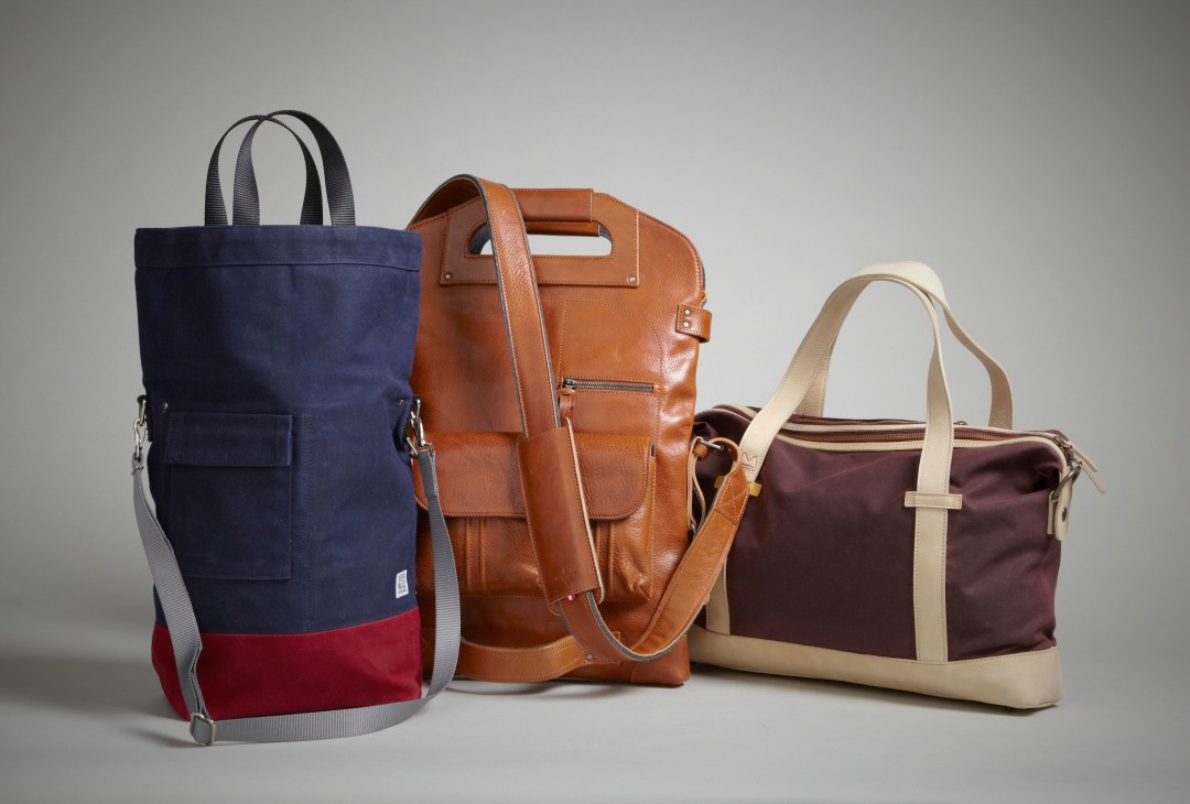 Three bags displayed: navy and red tote, cognac leather backpack, and burgundy with cream handles shoulder bag.
