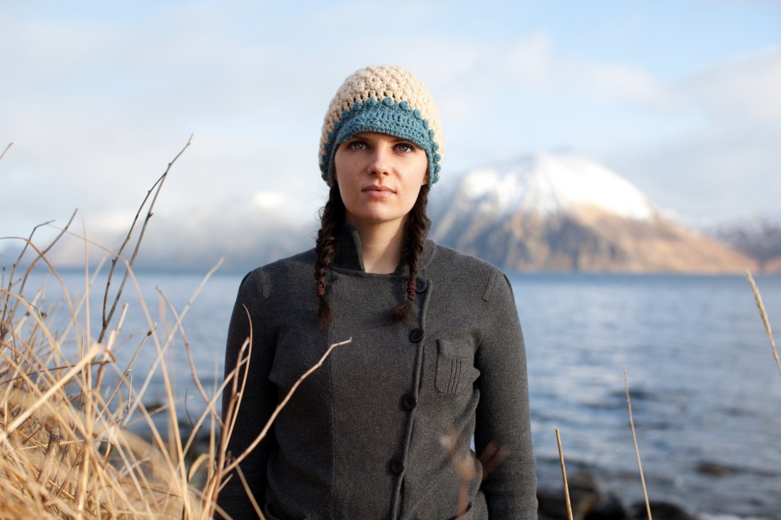 Woman wearing a cream and blue knitted hat standing by the Aleutian coast with snow-capped mountains in the background.
