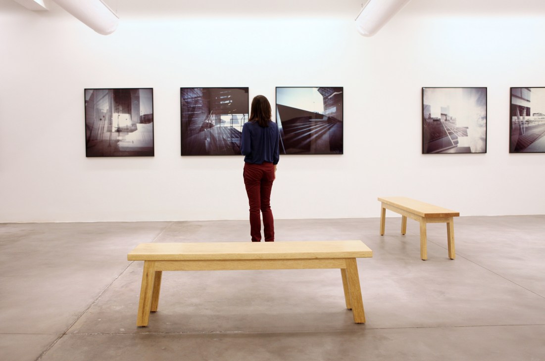 Visitor viewing framed architectural photographs in a minimalist white-walled gallery space with wooden benches.