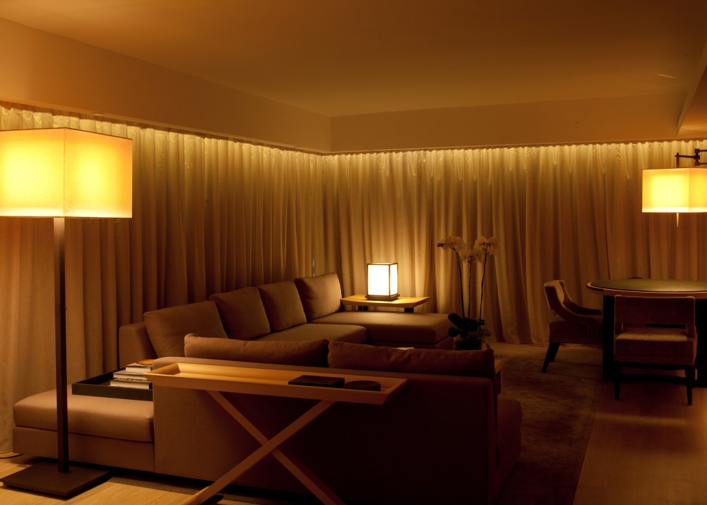 Warm-lit hotel suite with cream sofa, floor lamps, and floor-to-ceiling curtains at The Upper House.
