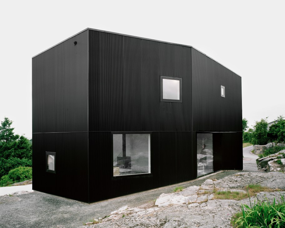 Black corrugated metal house on hilltop overlooking archipelago with large windows and minimal openings.