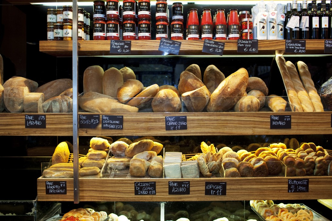 Doppio Zero bakery display window with fresh breads and pastries on wooden shelves and bottled goods above.