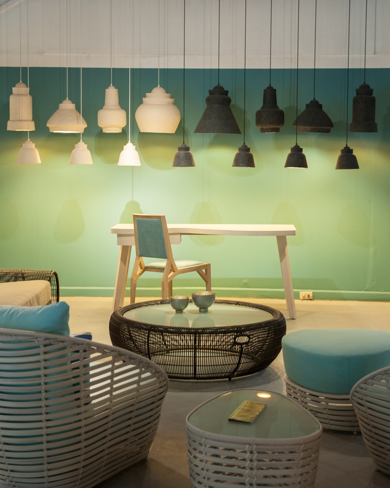 Modern showroom display featuring hanging pendant lamps in cream and black, wooden table, woven chairs and round table with cushions against a green wall.