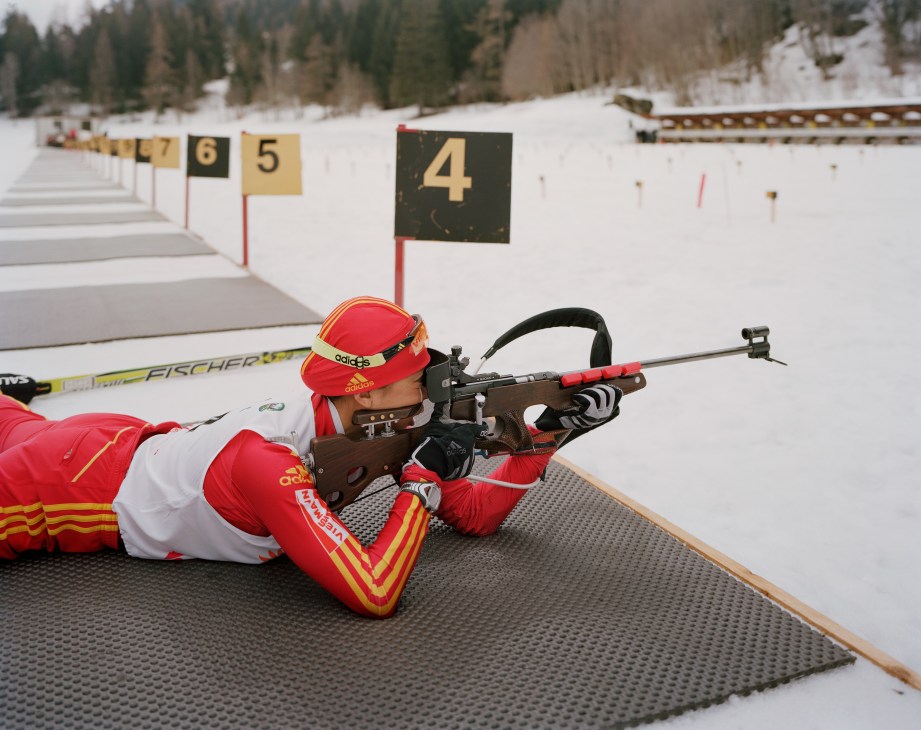 Military biathlon competitor in red uniform aiming rifle at snowy shooting range with numbered targets.