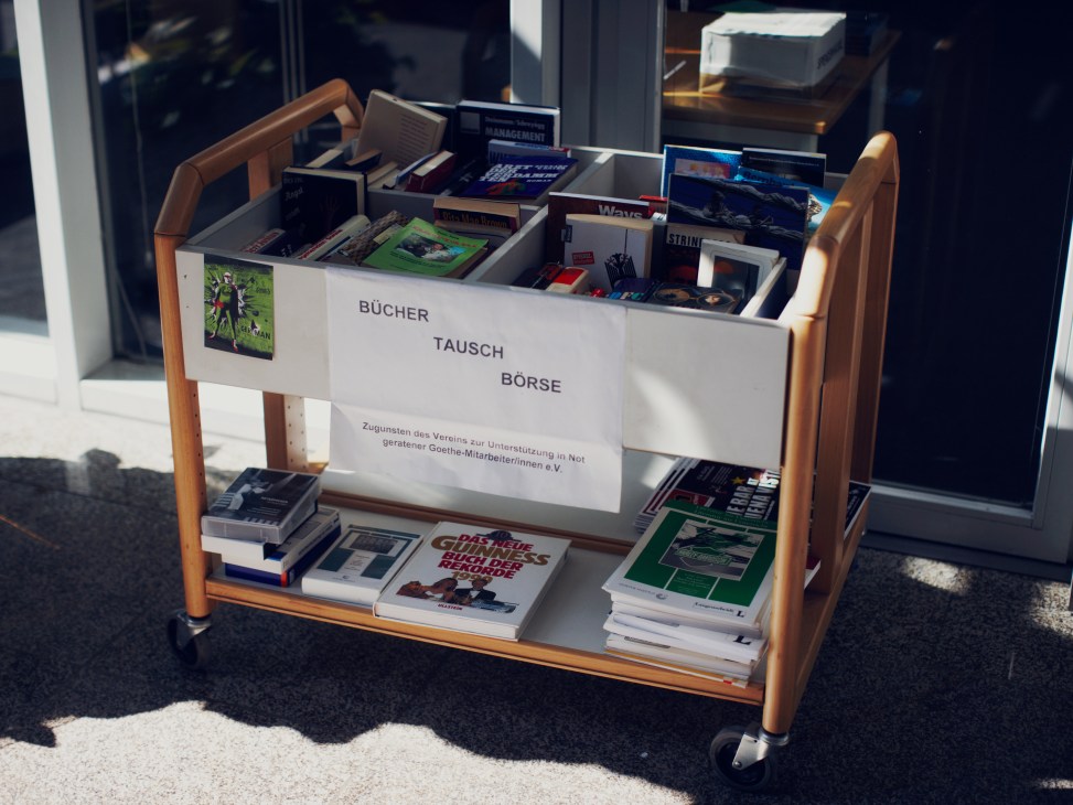 A wooden cart displaying books and materials labeled 'Bücher Tausch Börse' (book exchange) for Goethe-Institut employees.