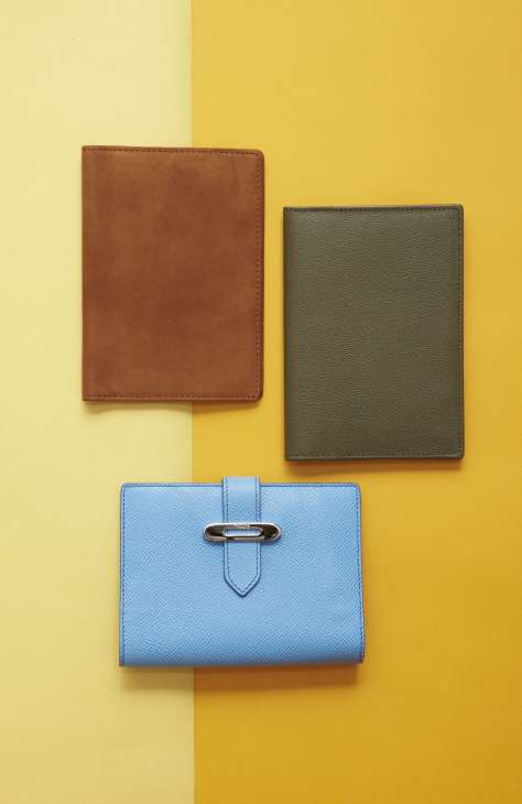 Three leather passport cases in cognac, olive green, and light blue arranged on a split yellow background.