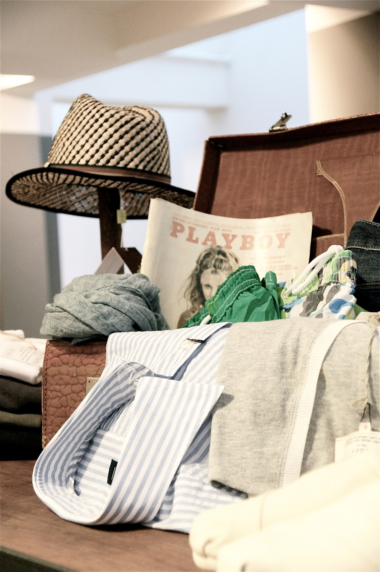 Feldenkirchen retail display with clothing, a straw hat, brown leather bag, and Playboy magazine.