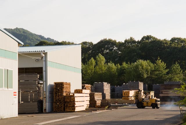 Stacks of lumber and wood materials stored outside a Maruni factory building with forested hills in the background.