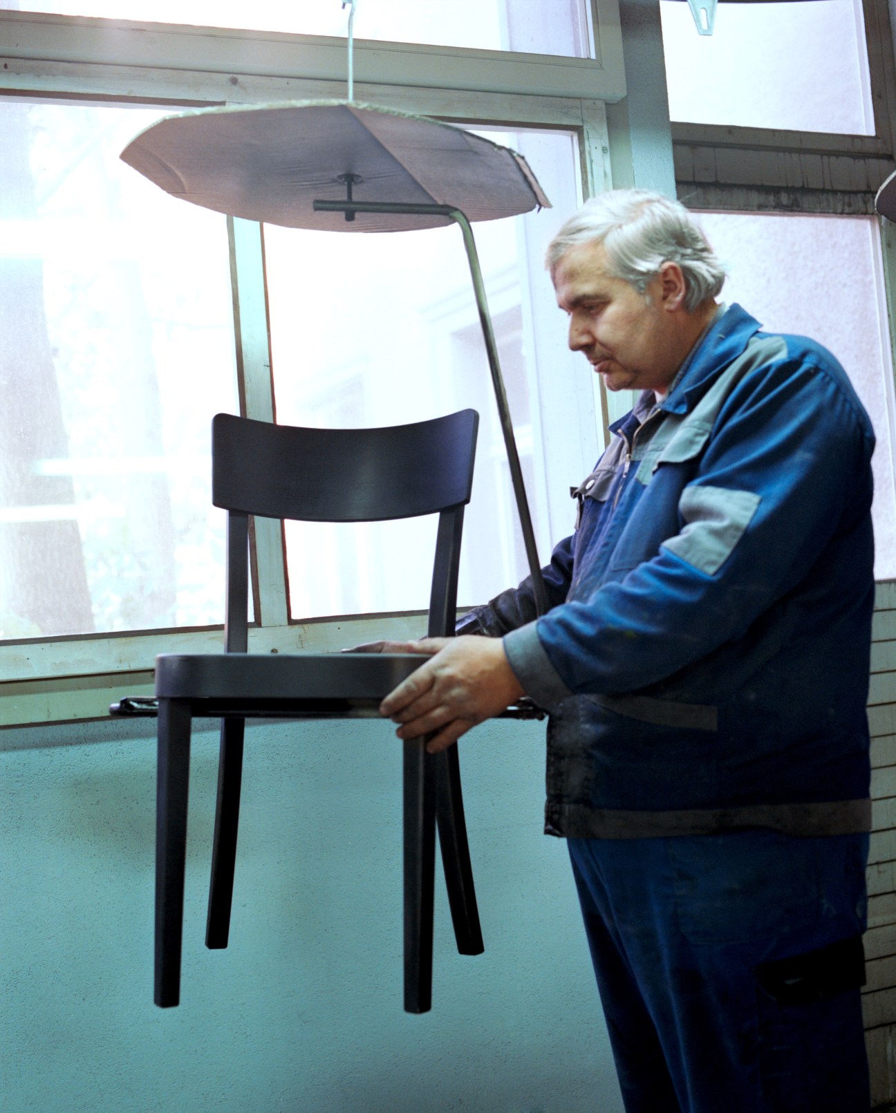 Worker in blue jacket applying varnish to a black wooden chair in the paint shop.