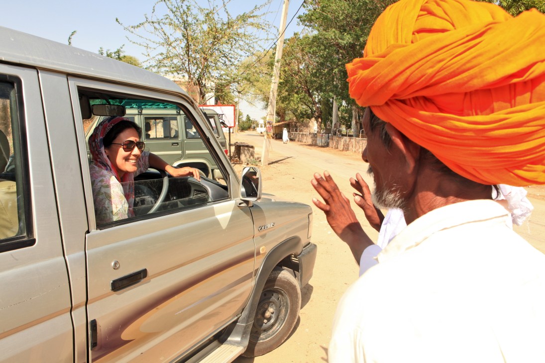 A woman in sunglasses and headscarf drives a beige SUV past a man wearing a bright orange turban on a rural dirt road in Soda village.