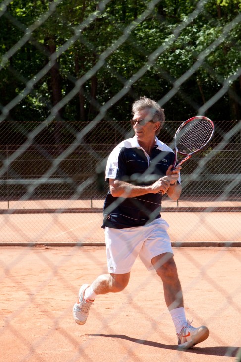 An older man plays tennis on a clay court in Paris, wearing a navy and white polo shirt.