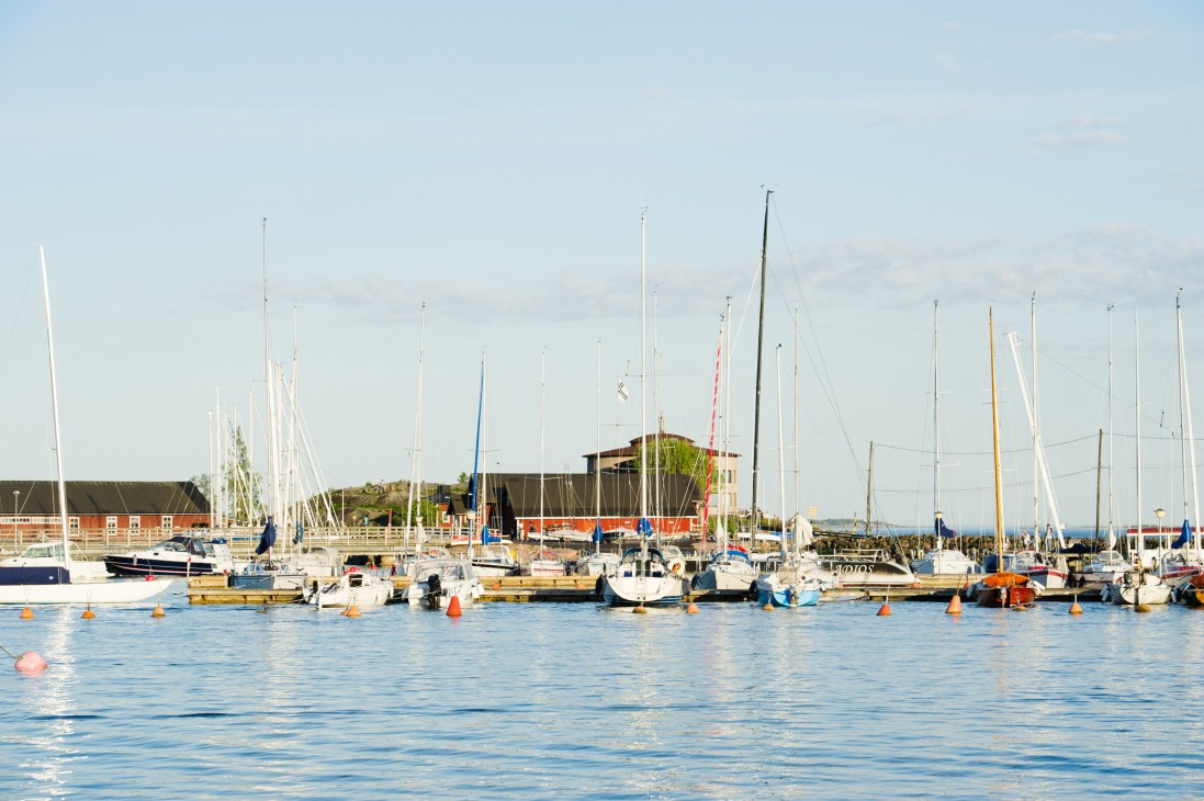 Sailboats moored in a marina with waterfront buildings and clear sky.