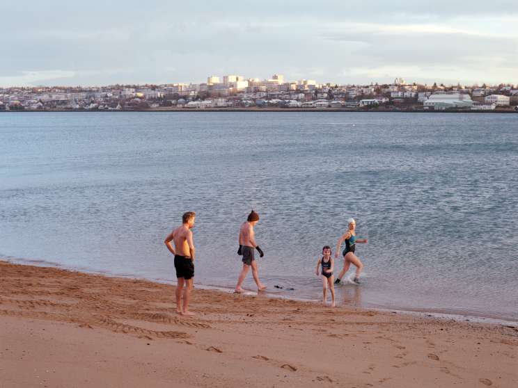 People wading into cold seawater at a Reykjavík beach with the city skyline visible across the bay.