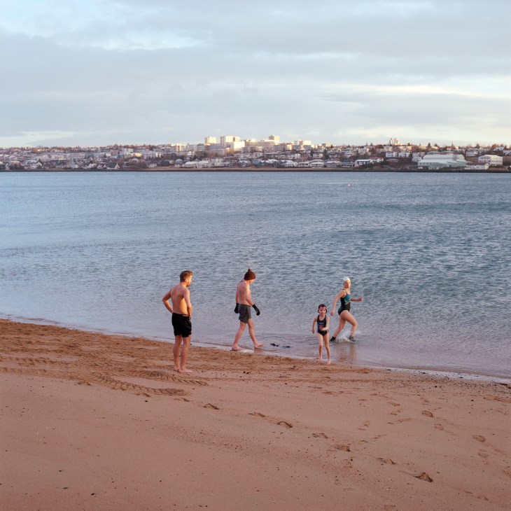People wading into cold seawater at a Reykjavík beach with the city skyline visible across the bay.