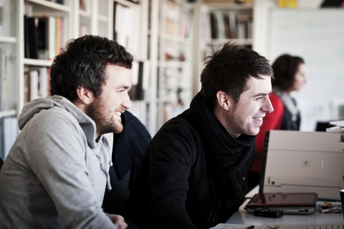 Two men smiling while working at computers in a bright office with bookshelves in the background.