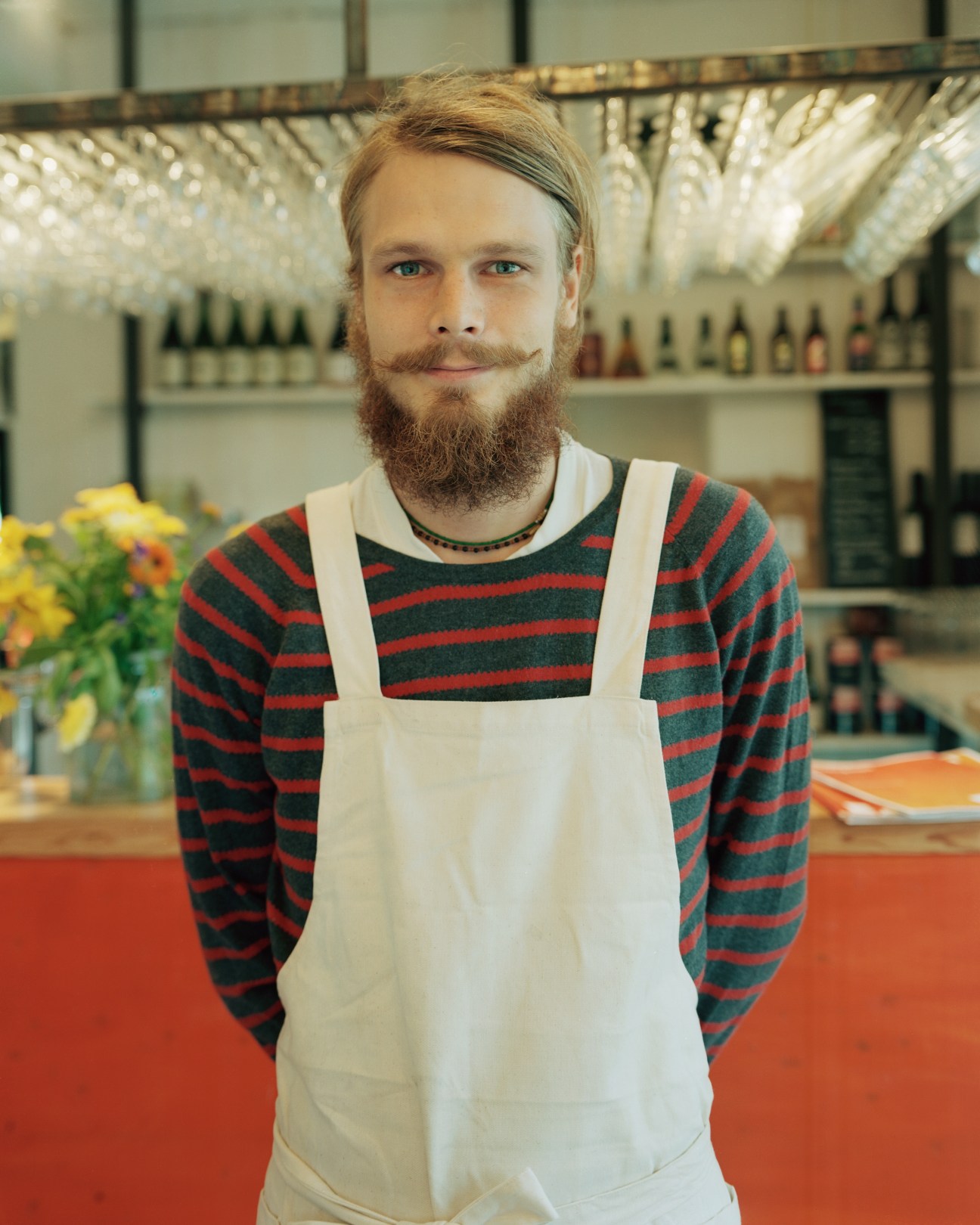 Petter Söderhäll, a bearded waiter wearing a striped shirt and white apron, stands in front of shelves of bottles at Leva café/restaurant.