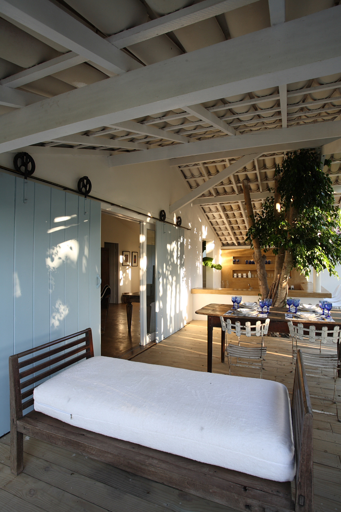 Interior of Jacaré do Brasil villa showing bedroom with wooden bed, blue doors, and open-plan living area with dining table.