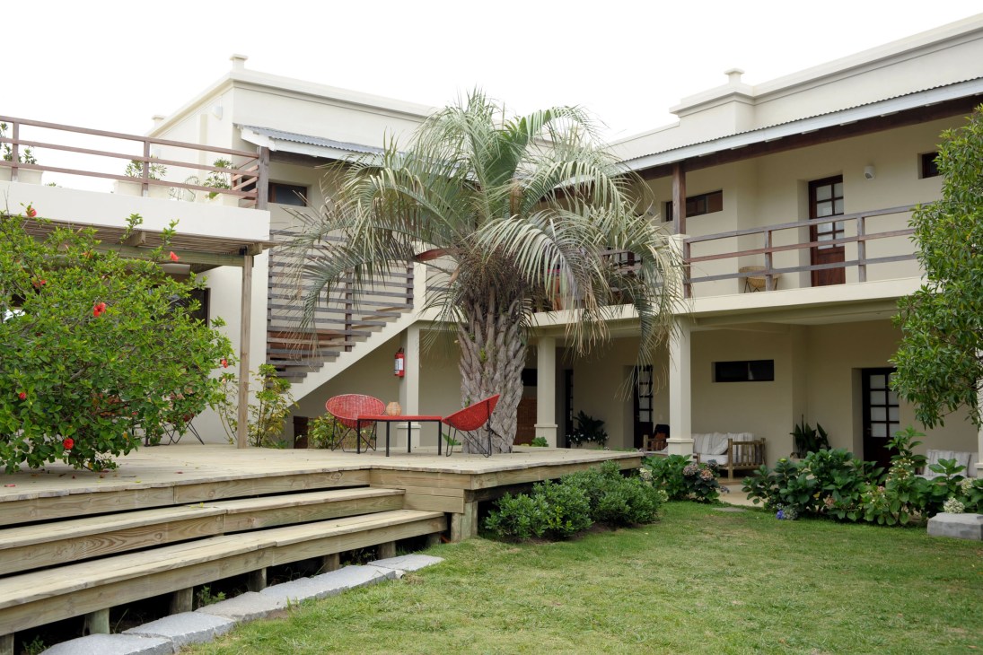 Modern multi-level house with white walls, palm tree, red chairs, and wooden deck at Brisas de la Pedrera.