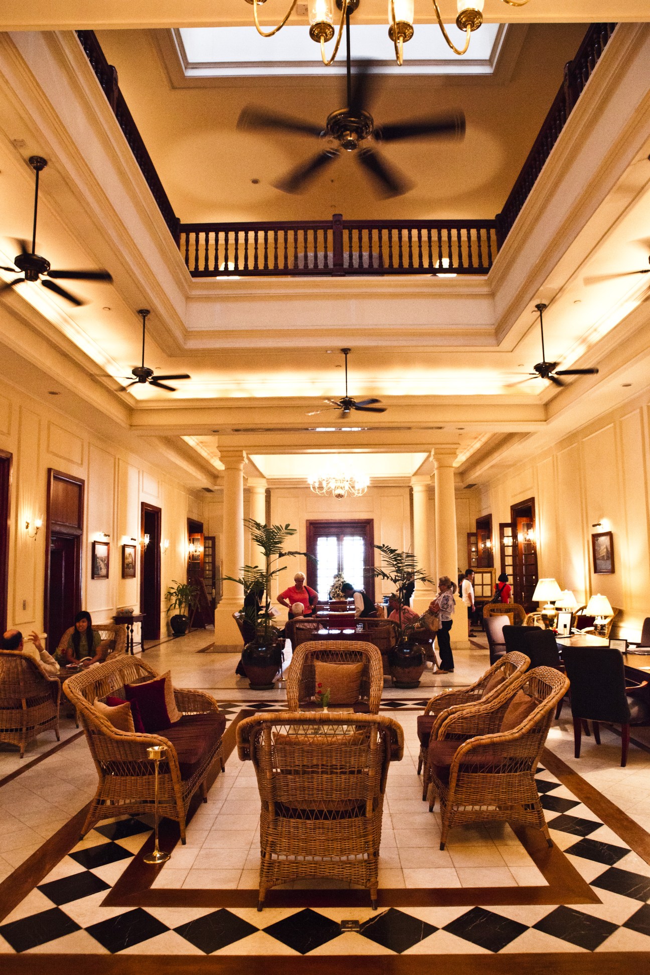 The Strand Hotel's elegant colonial lobby with high ceiling, brass chandeliers, wicker chairs, and black-and-white checkered floor.