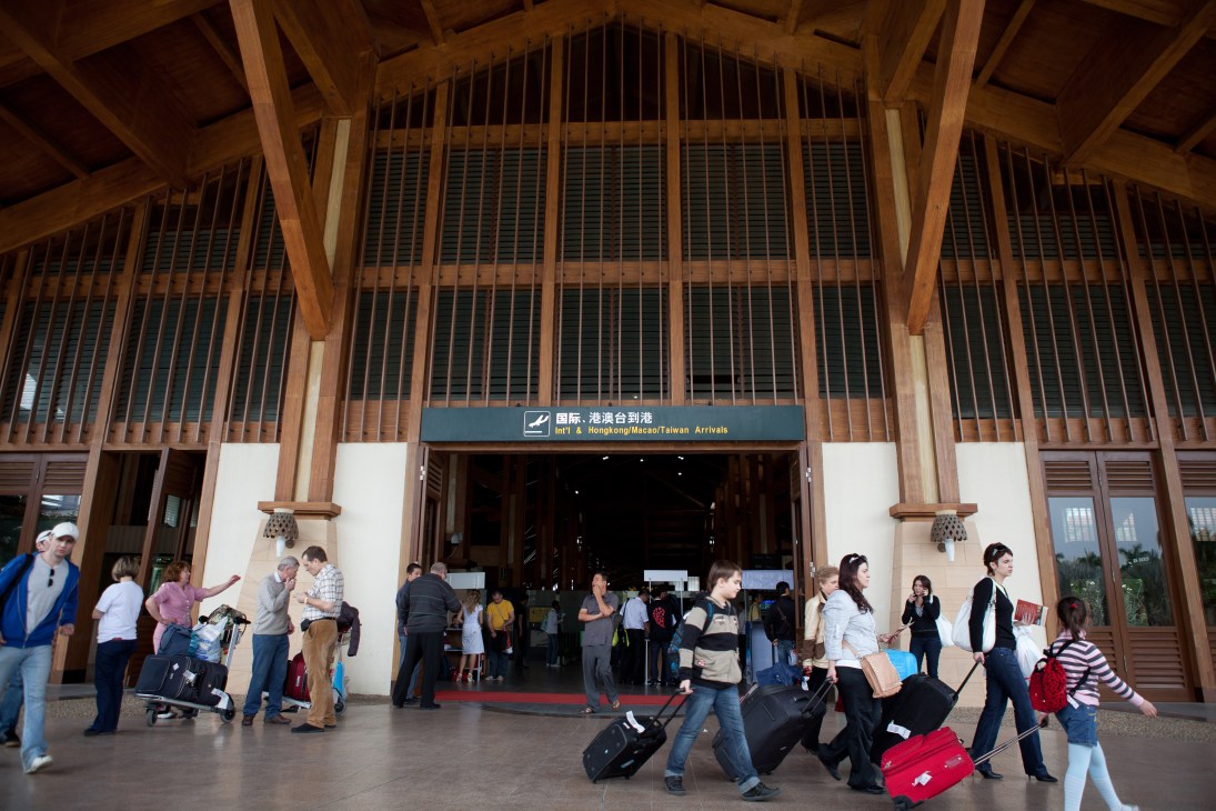 Passengers with luggage in the arrivals hall at Sanya Phoenix International Airport with wooden ceiling and bilingual signage.