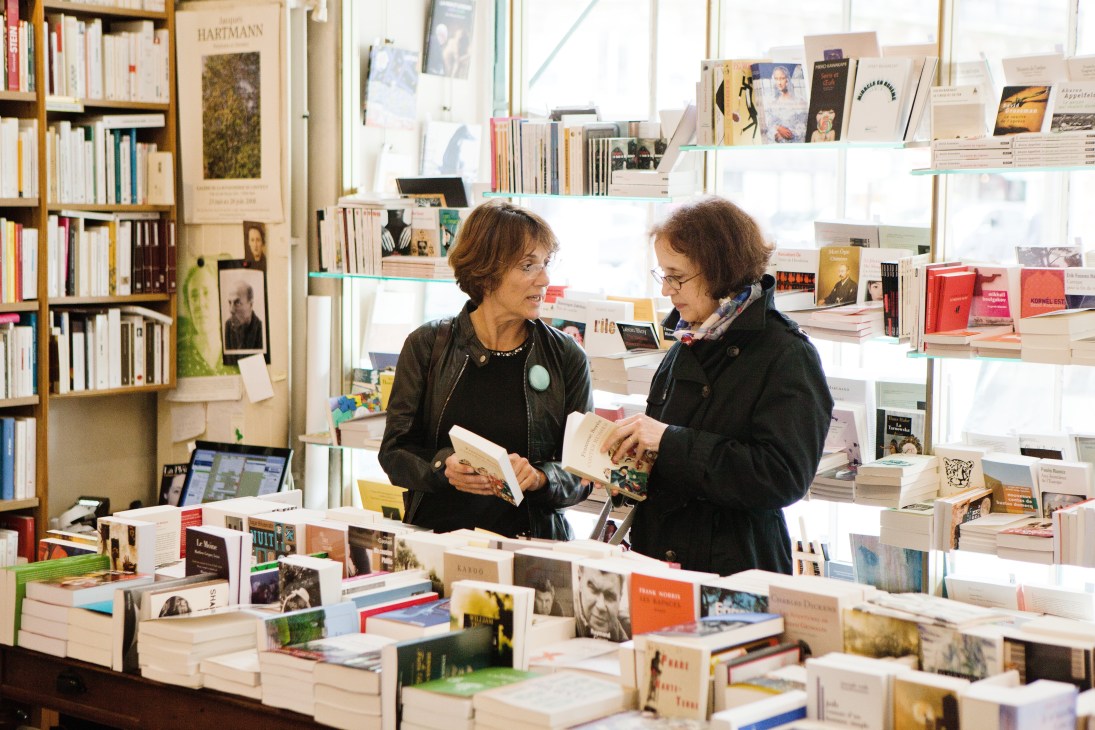 Two women browse and discuss books at Tschann Libraire, a Paris bookshop with white shelving and wooden displays.