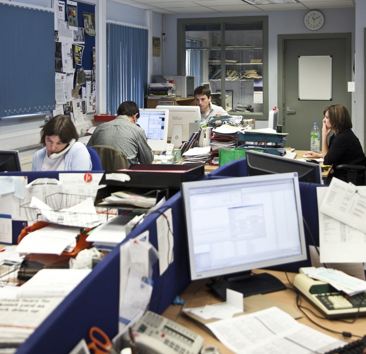 Newsroom staff working at desks with computers and papers in the West Highland Free Press office.