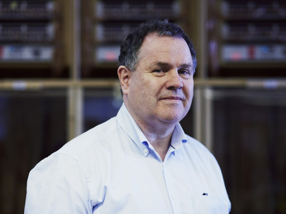 Charlie Brennan, CEO of Arcam, wearing a light blue shirt in front of audio equipment.