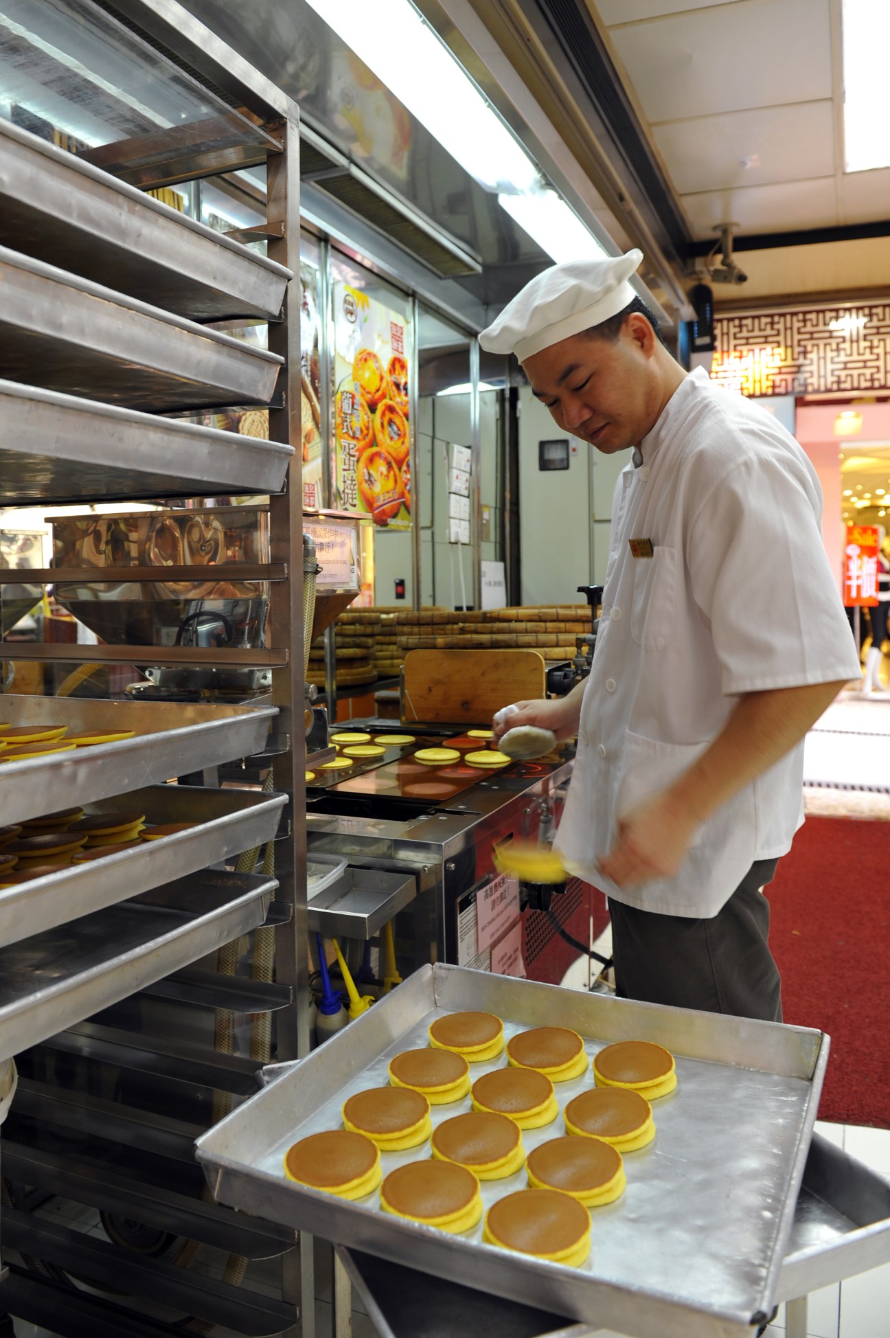 Baker in white chef's hat preparing egg tarts in a Macau kitchen with industrial ovens.