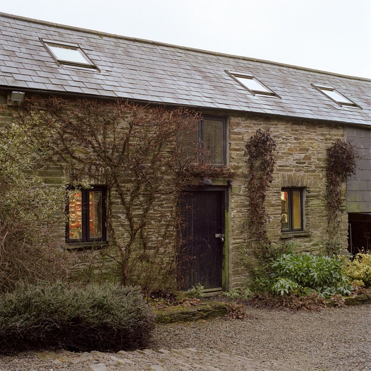 Stone farmhouse with slate roof, dark windows and door, surrounded by bare vegetation.